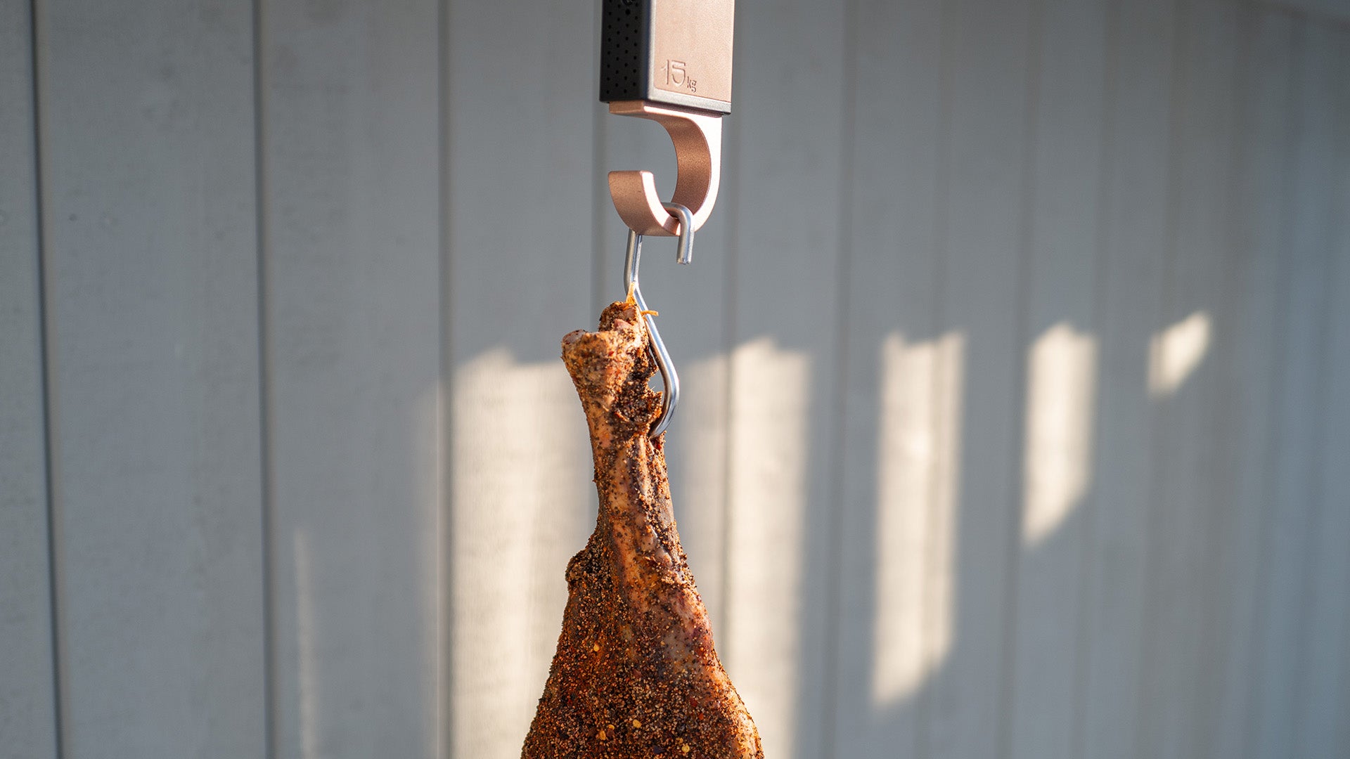 Smoked leg of cured meat hanging from a hook against a gray wall.