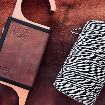 Copper meatdryer with leather handle on a brown surface next to black and white twine, string