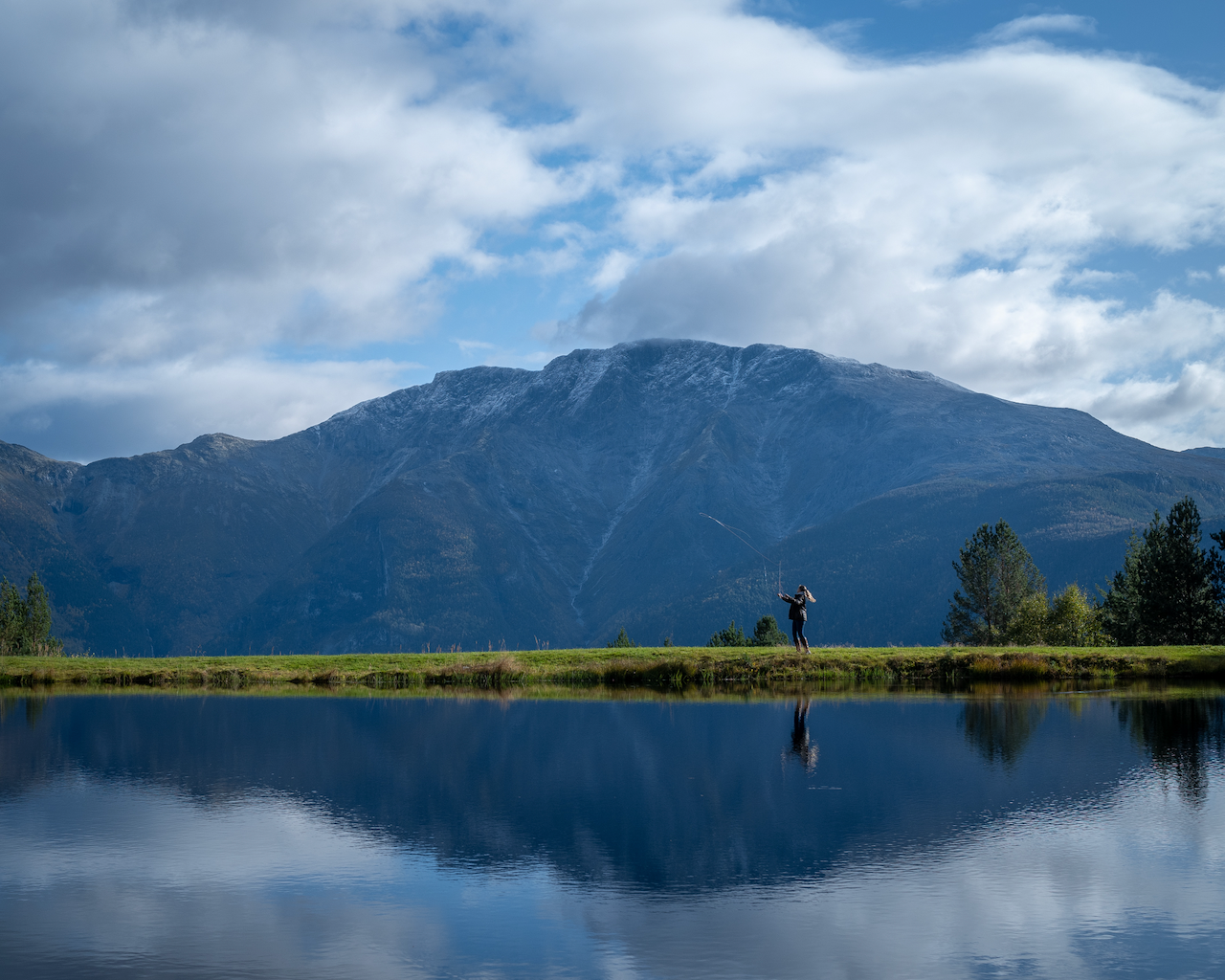 Woman standing on a grassy area by a lake , fly fishing with mountains in the background